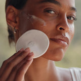 Woman applying a white round compact to her face with a blurred natural background