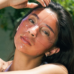 Woman applying sunscreen to her face outdoors with greenery in the background