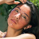 Woman applying sunscreen to her face outdoors with greenery in the background