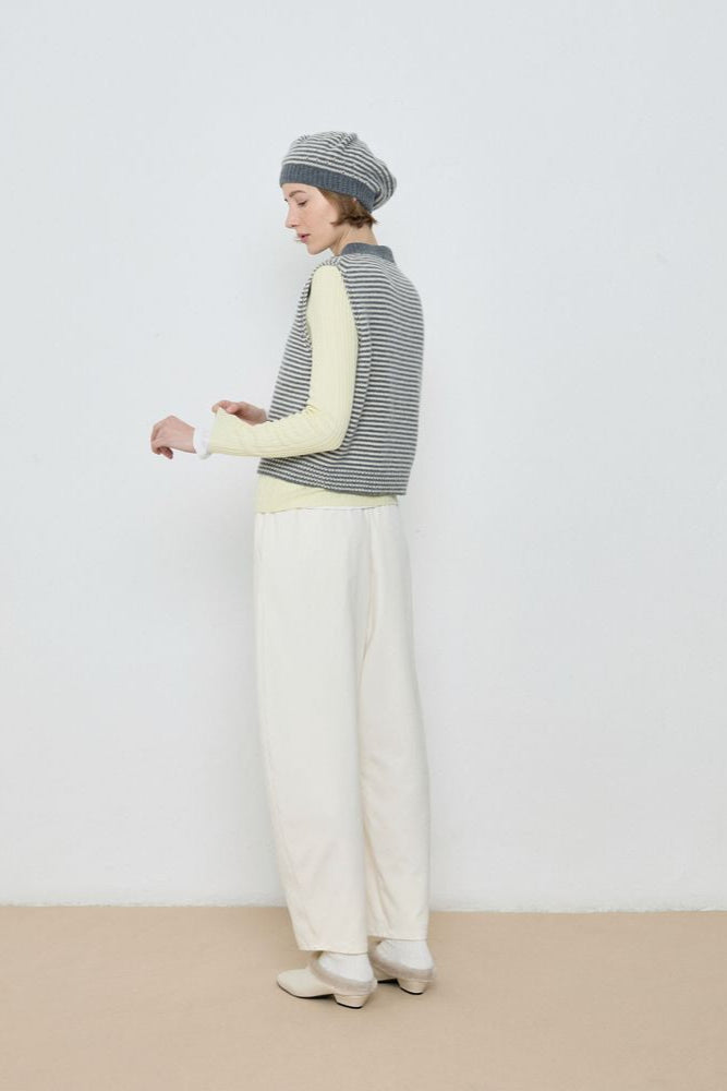 Person wearing a striped sweater vest, white pants, and a beanie against a plain background