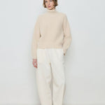 Person wearing a beige sweater and white pants against a plain background