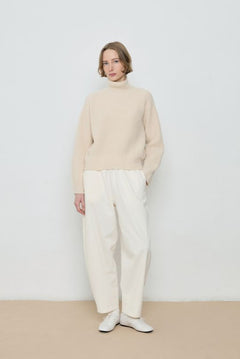 Person wearing a beige sweater and white pants against a plain background