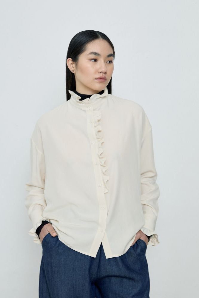 Woman wearing a white blouse with ruffled details and dark pants against a plain background