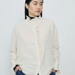 Woman wearing a white blouse with ruffled details and dark pants against a plain background