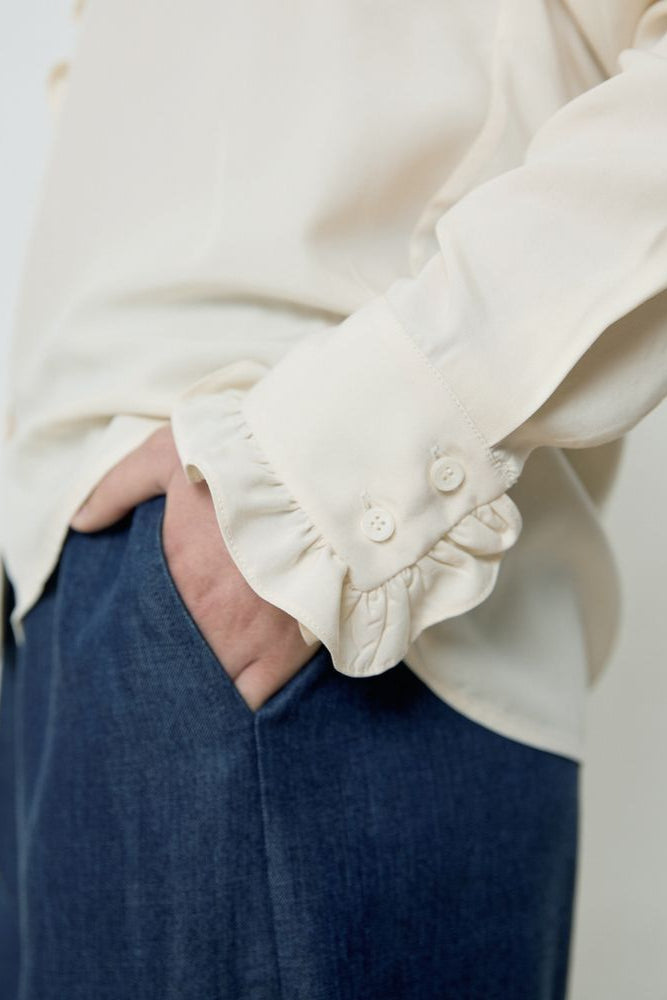 Person wearing a white blouse with ruffled cuffs and blue pants on a plain background
